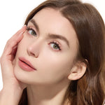 Close-up of a woman with brown hair and green eyes, touching her face.